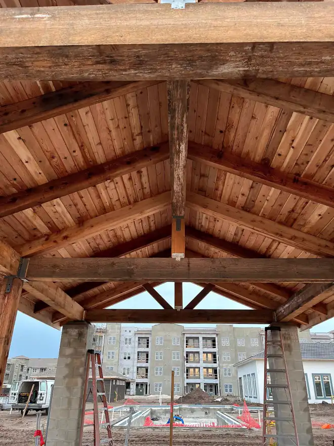 Finished cedar pavilion with heavy timber posts and exposed roof joinery, apartment complex beyond